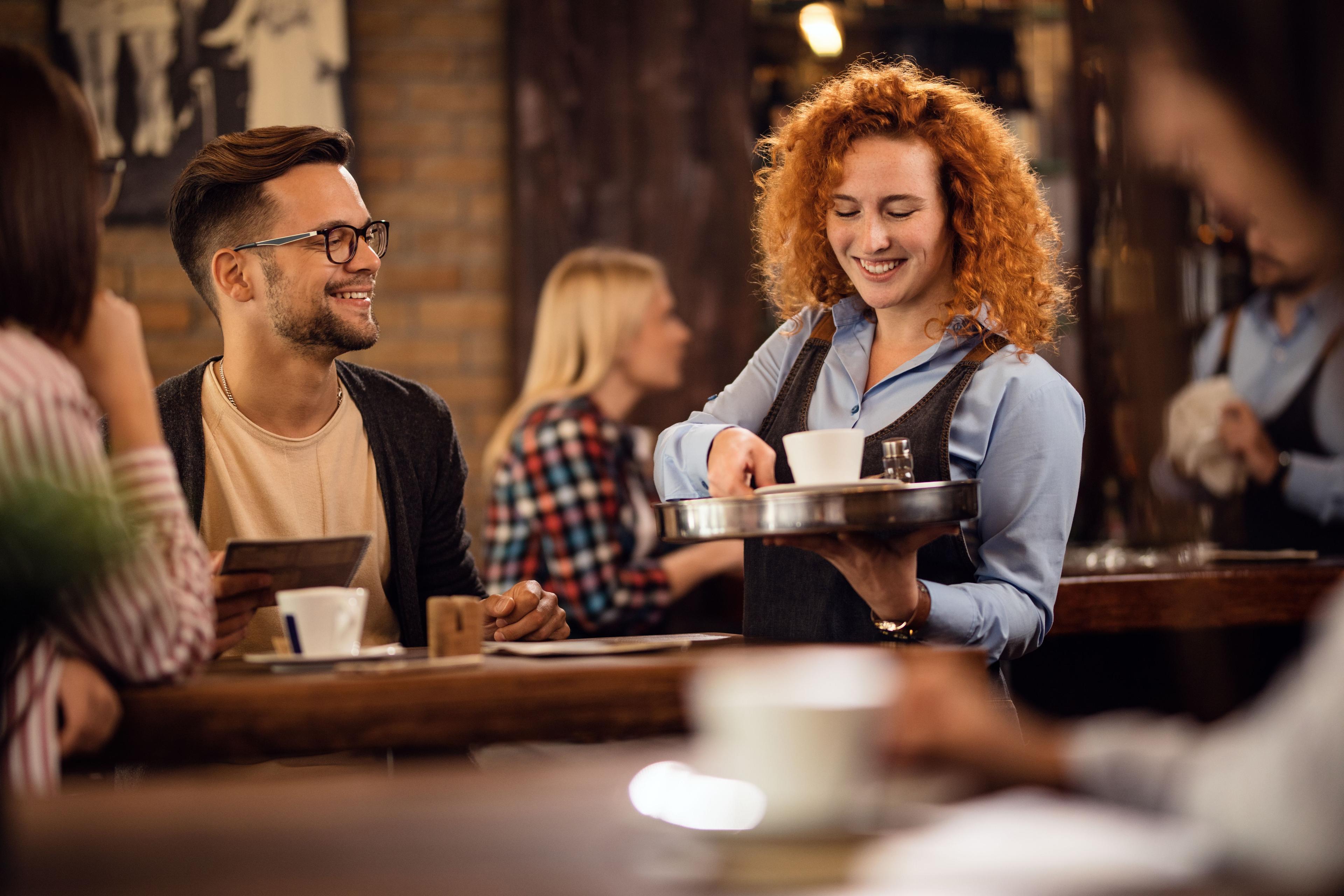 A smiling waitress serves coffee to customers at a cozy, dimly lit cafe.