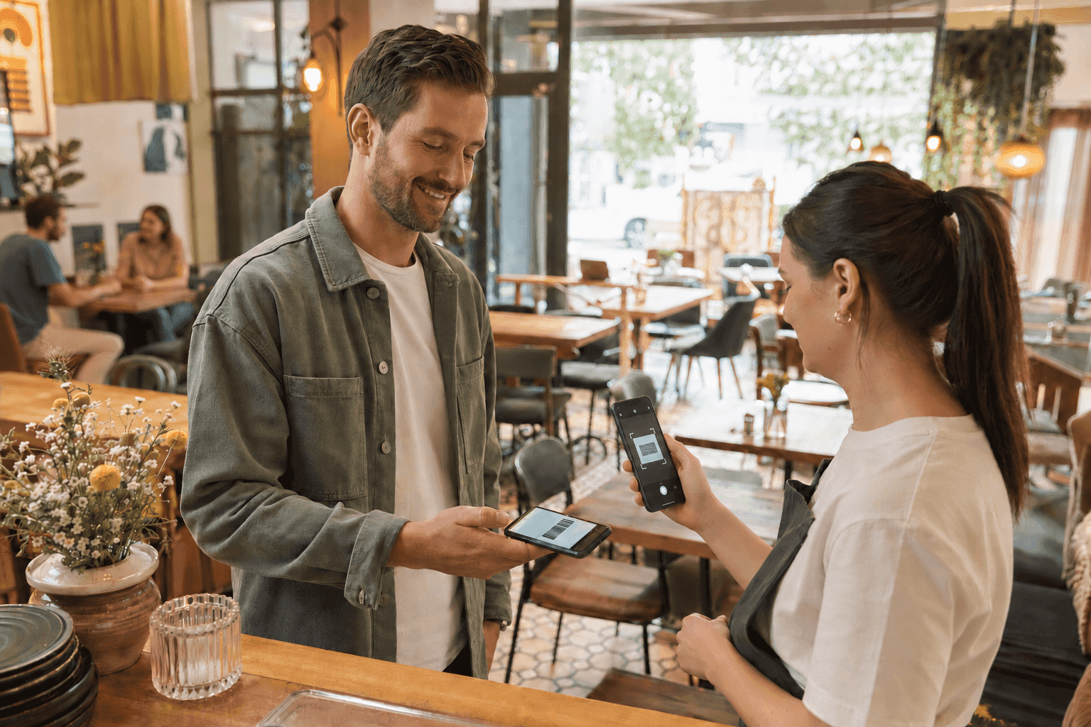 Man uses his smartphone for contactless payment at a cafe counter with a smiling cashier.