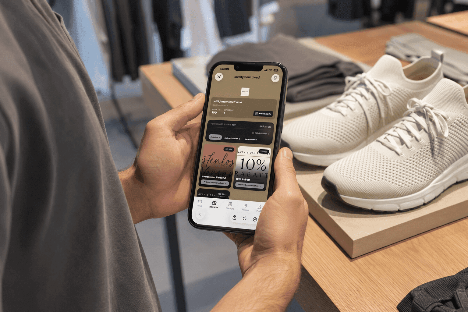 Hands hold a smartphone displaying a shopping app next to pairs of white sneakers.